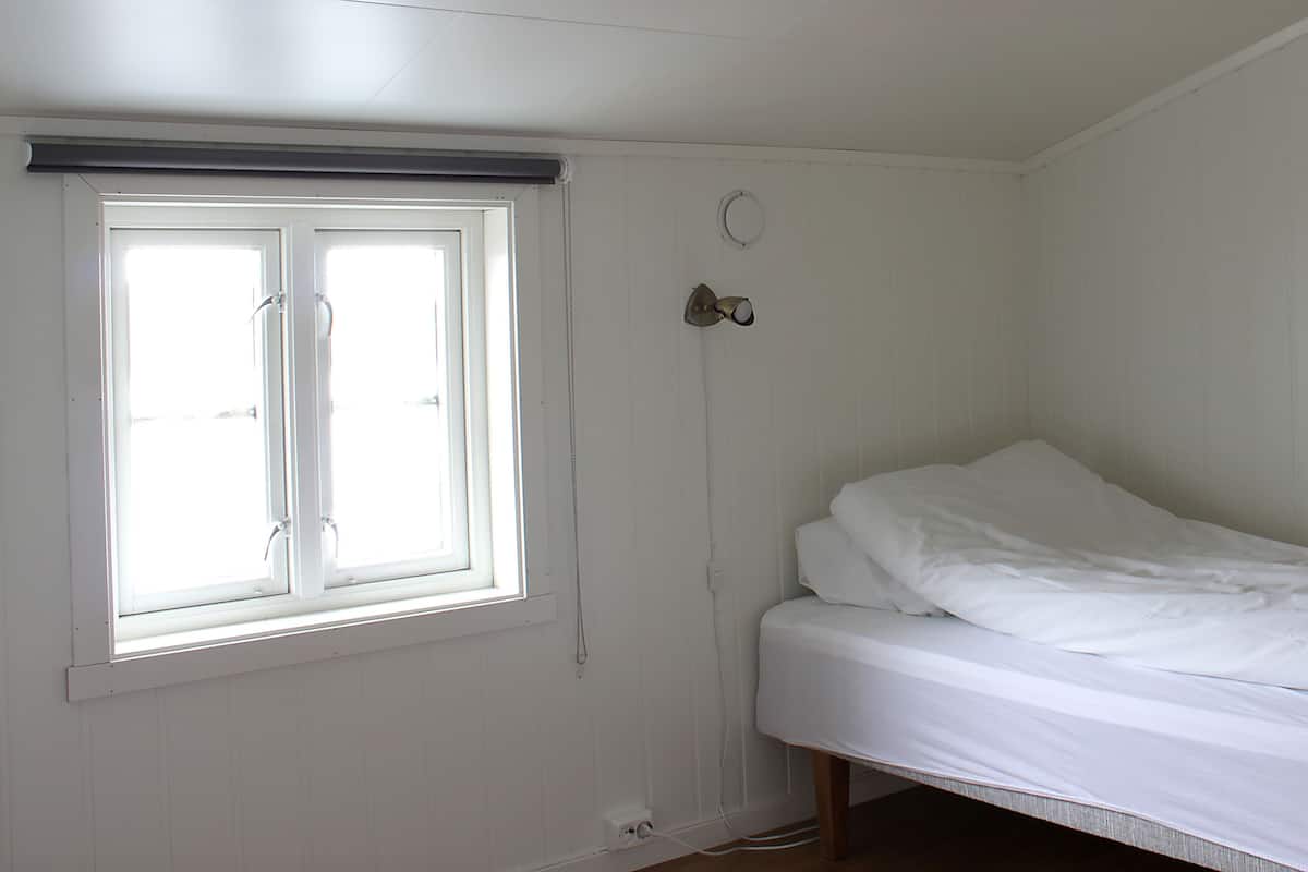 A small, white room with a single bed, a window, and a wall-mounted light fixture.