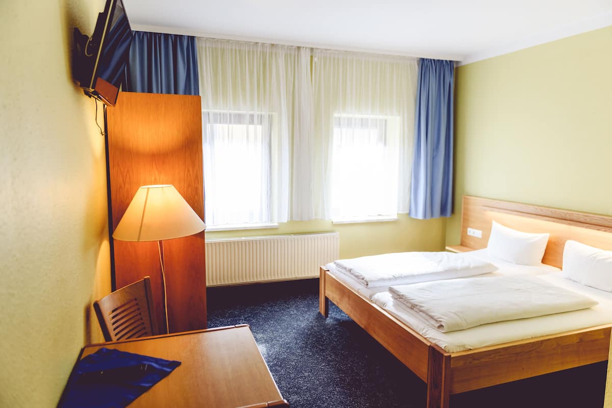 Une chambre d’hôtel avec un lit en bois, un bureau, une chaise, une lampe et une fenêtre avec des rideaux.