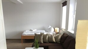 A modern, compact living space with a wooden floor, a brown sofa, a white side table with a plant, a bed with white bedding, and a window with blinds. - Landgasthof Schwabhausen (Schwabhausen)