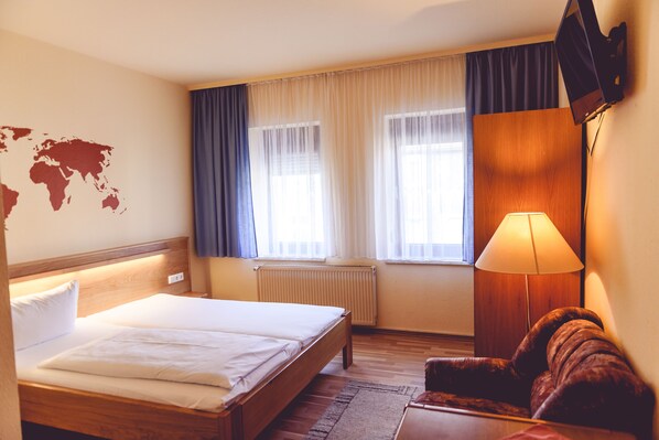 A hotel room with a bed, a wooden headboard, a world map on the wall, a window with curtains, a radiator, a wooden wardrobe, and a lamp.