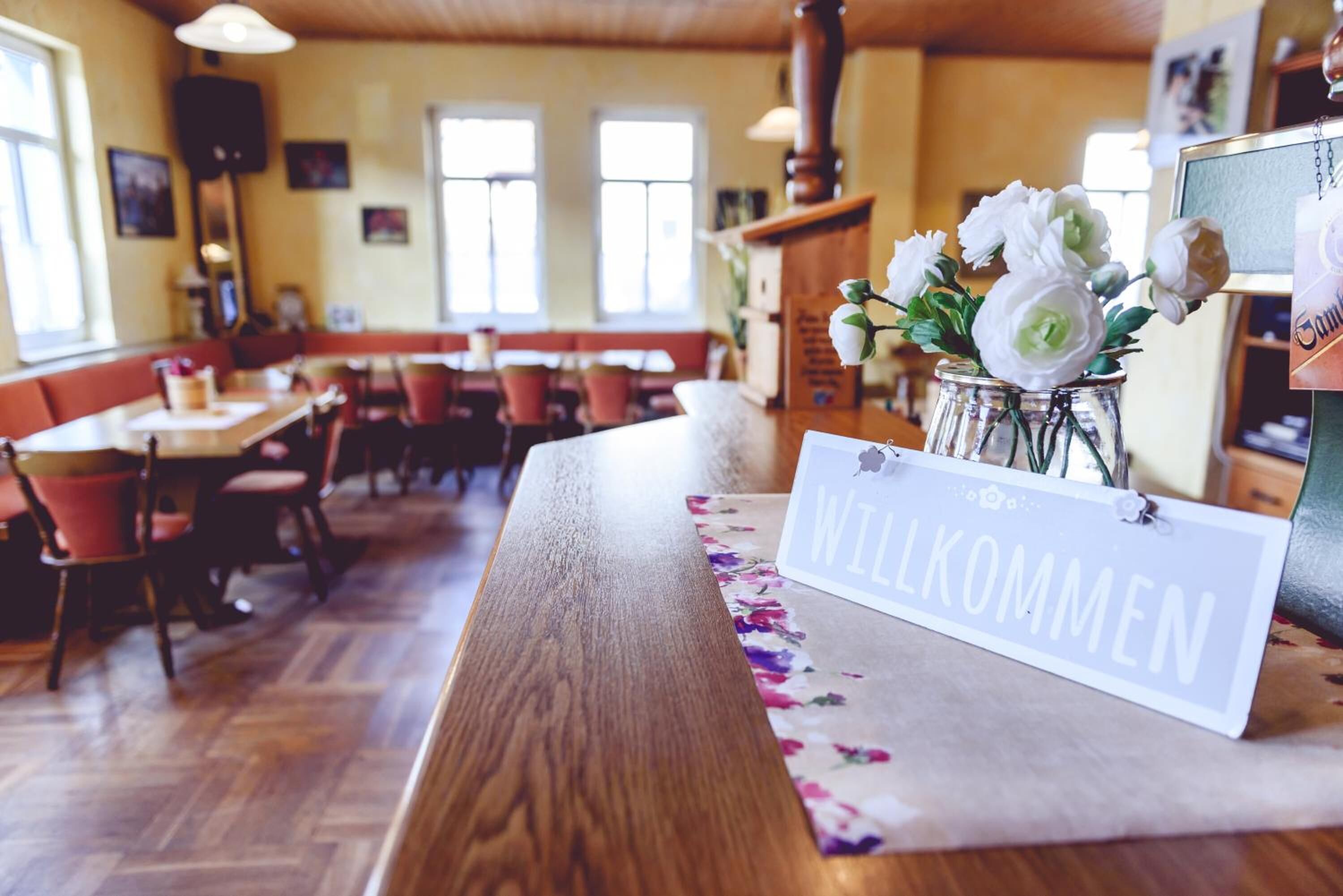 A restaurant interior with wooden tables and chairs, a 'Willkommen' sign, and a vase of white flowers.