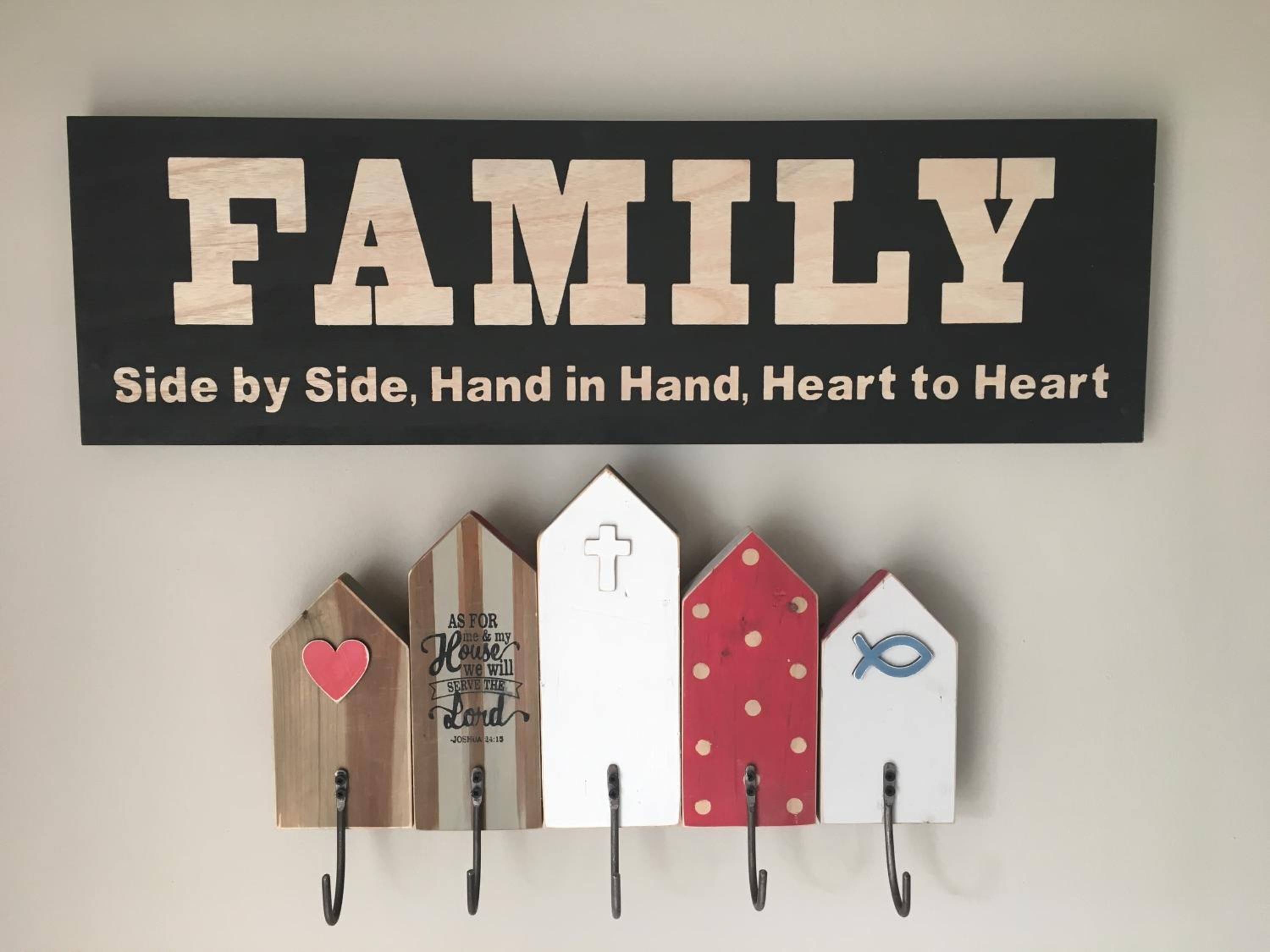 Et ophængt skilt med ordet 'FAMILY' og teksten 'Side om side, hånd i hånd, hjerte til hjerte' over en række træhuse-formede kroge med forskellige symboler og tekst.