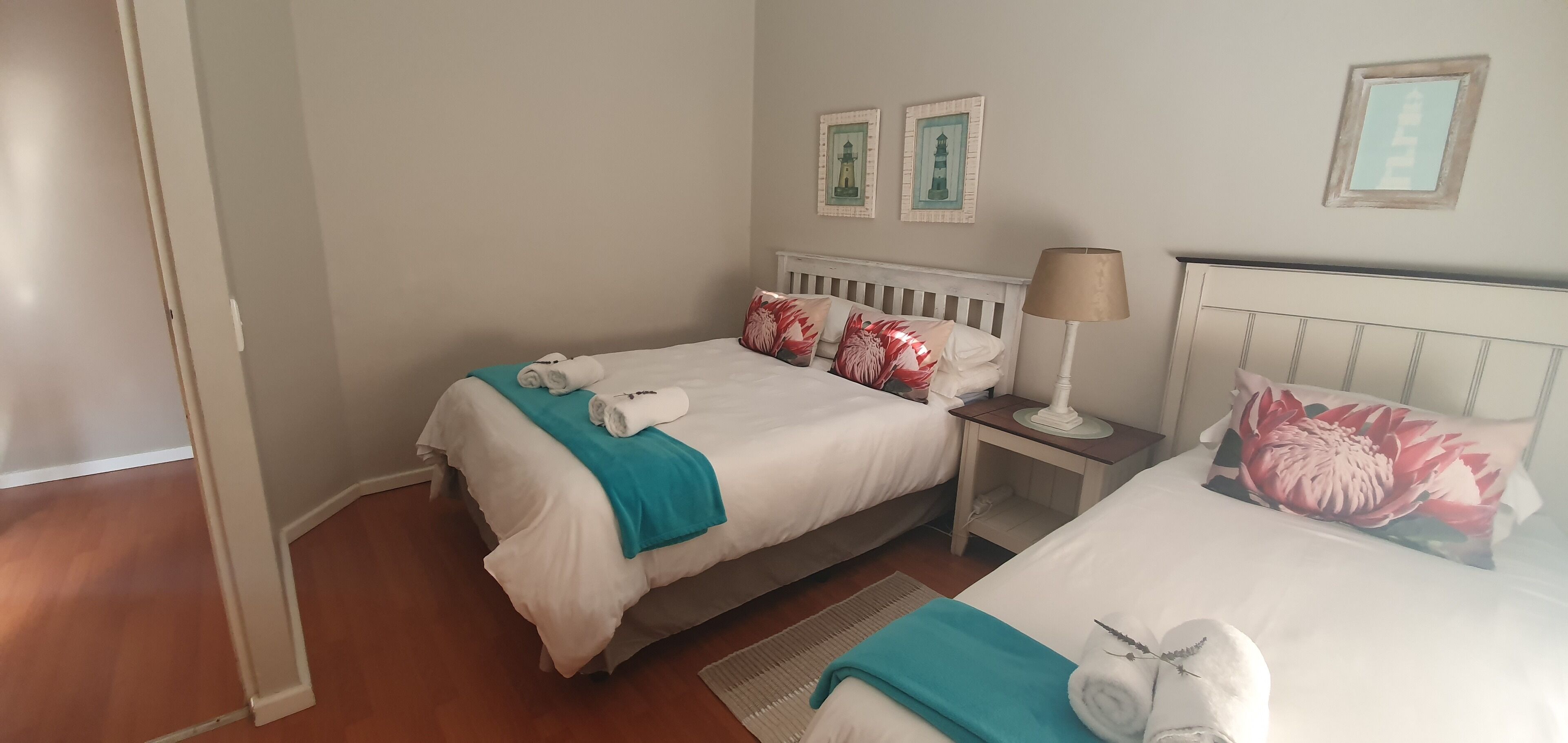 A hotel room with two beds, each with white bedding and teal throw blankets. There are decorative pillows, a bedside lamp, and framed artwork on the wall.