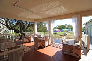 Un área de comedor al aire libre con techo, mobiliario de mimbre y vista a un parque infantil.