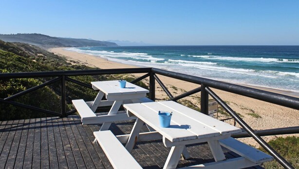 Een houten terras met picknicktafels en banken, met uitzicht op een strand met golven.