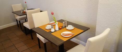 En spiseplads med et bord dækket til to personer, inklusiv tallerkener, bestik og en vase med en blomst.