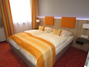 A hotel room with a large bed, orange and white striped bedding, a wooden headboard, two bedside tables with lamps, and a red patterned carpet. - Hotel Trutzpfaff (Speyer)