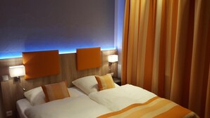 Habitación de hotel con dos camas, cada una con ropa blanca y detalles naranja, techo azul y cortina a rayas.