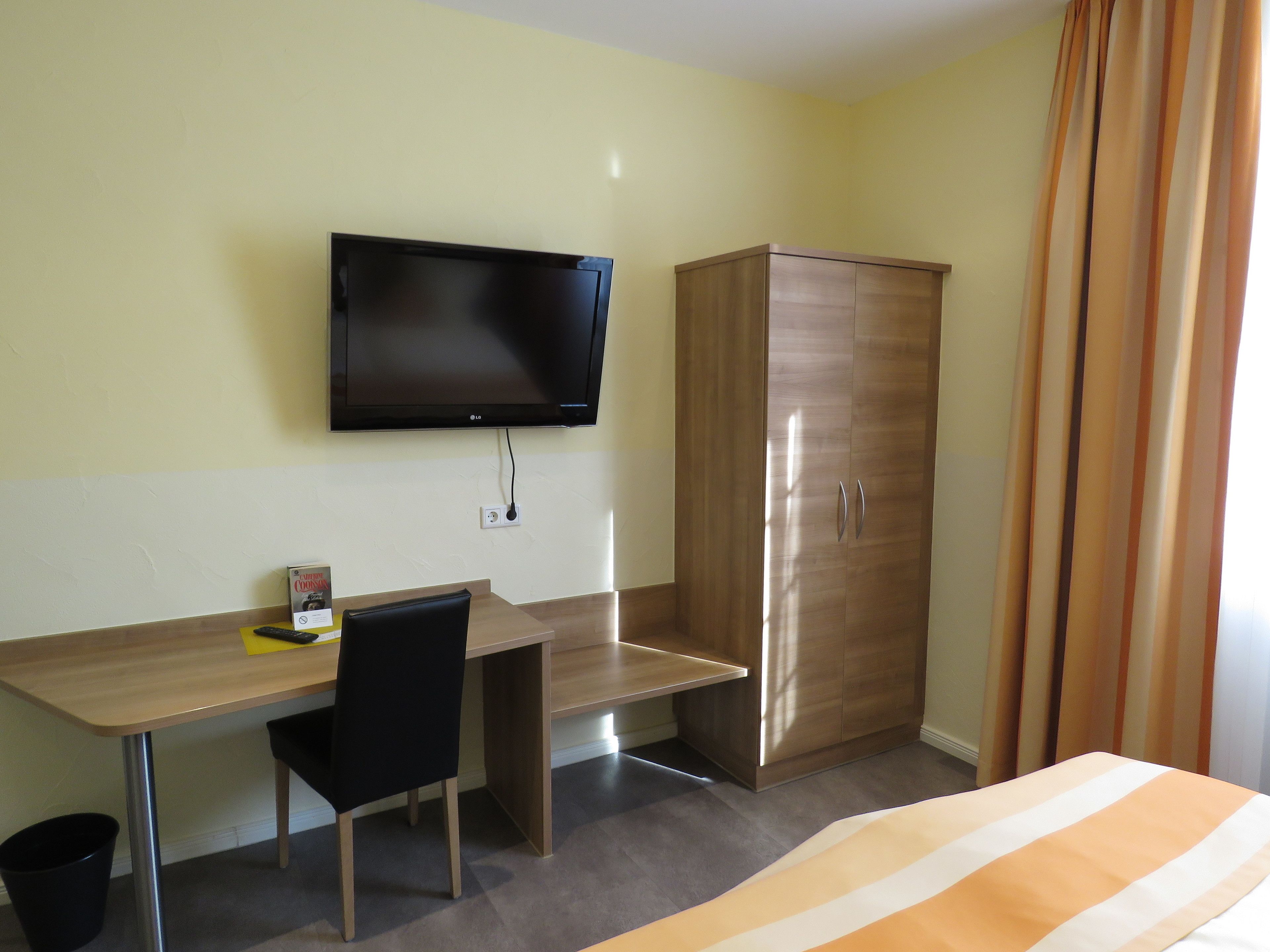 A hotel room with a wooden desk, a black chair, a television mounted on the wall, and a wardrobe.