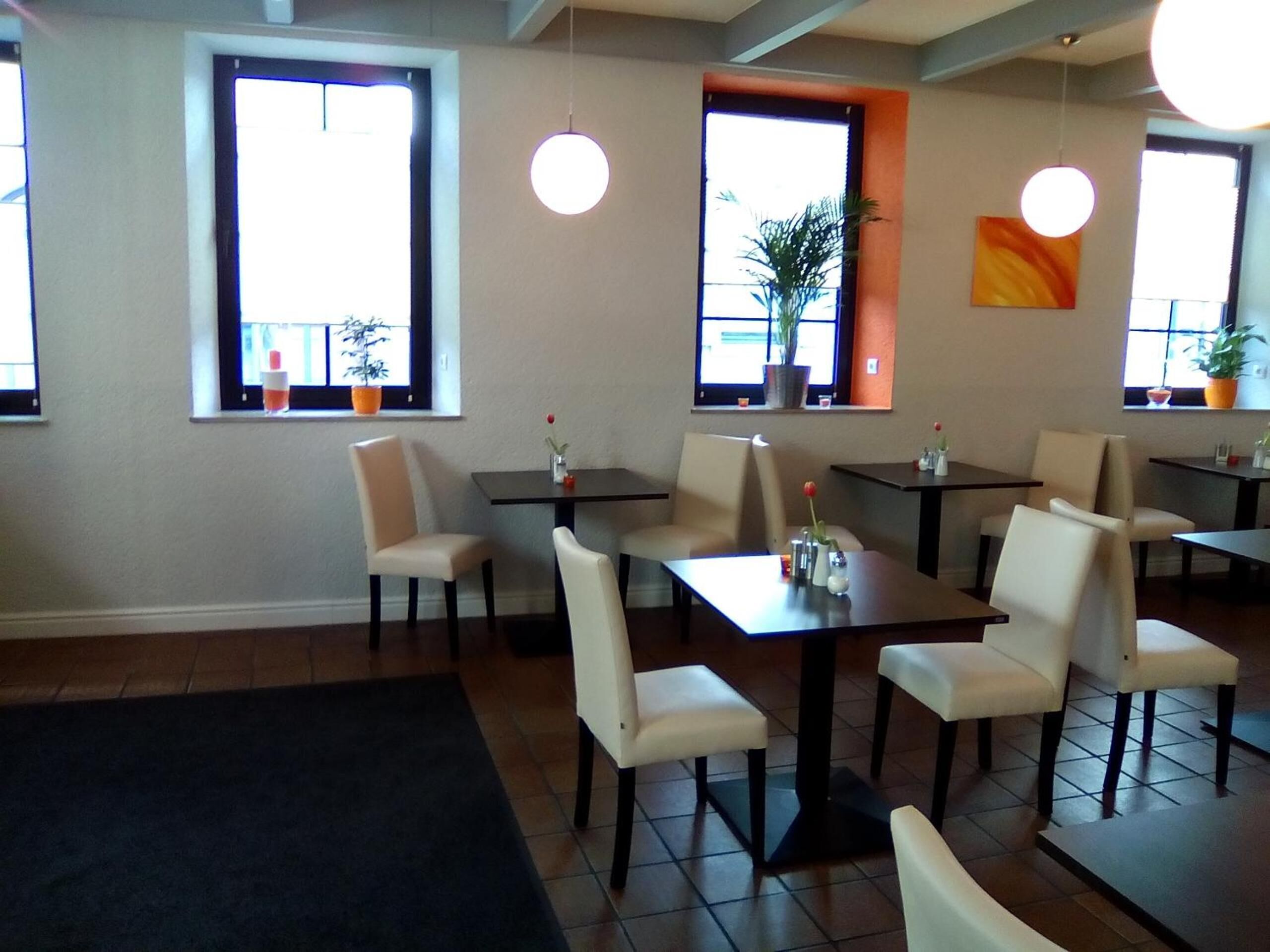 A restaurant with multiple tables and chairs, large windows with plants, and hanging pendant lights.