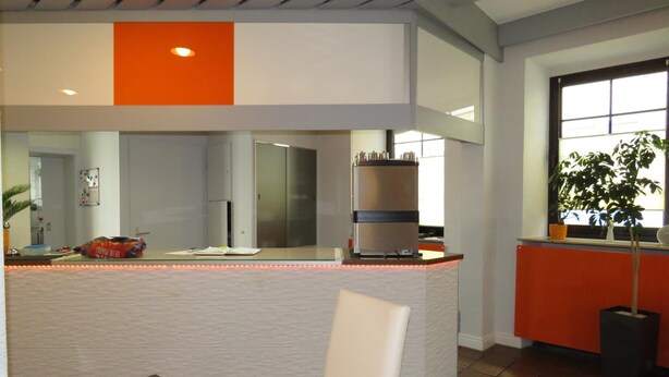 Una cocina moderna con una isla central, una zona de comedor con una computadora portátil sobre una mesa y un gran ventanal con plantas.