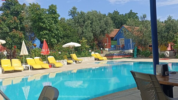 Une piscine entourée de bâtiments et d’arbres aux couleurs vives, avec des chaises longues jaunes et des parasols.