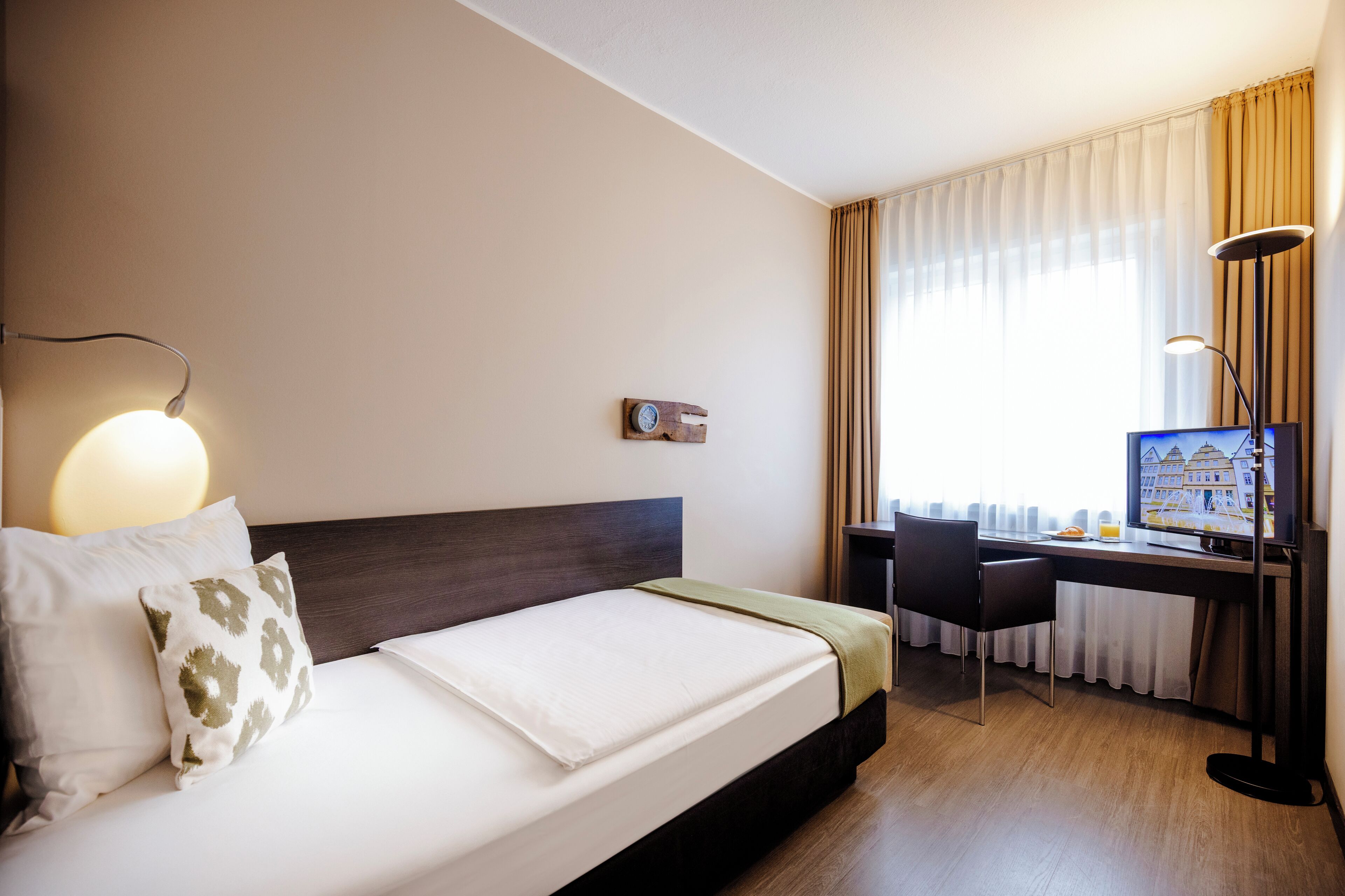 A hotel room with a bed, a desk, a chair, a lamp, and a window with curtains.