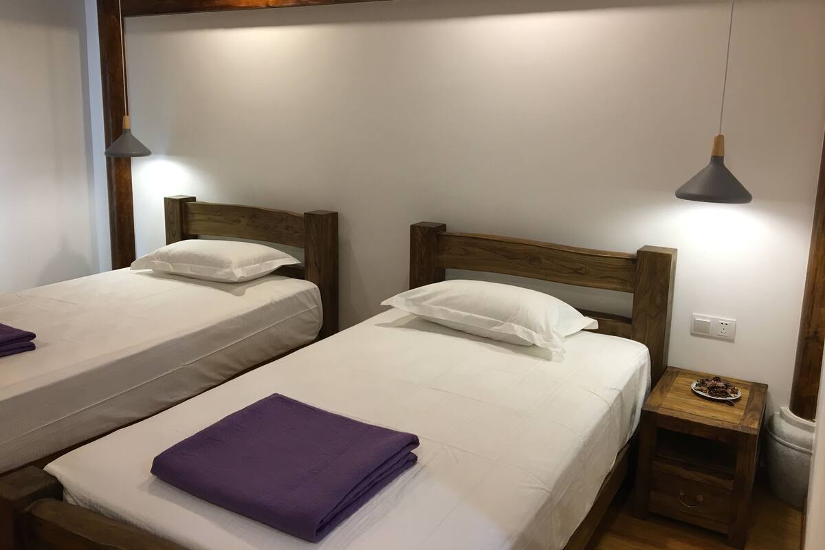 A room with two single beds, each with a wooden headboard, white bedding, and a purple blanket on one bed. There are two pendant lights hanging from the ceiling, and a small wooden nightstand with a plate of food on it.