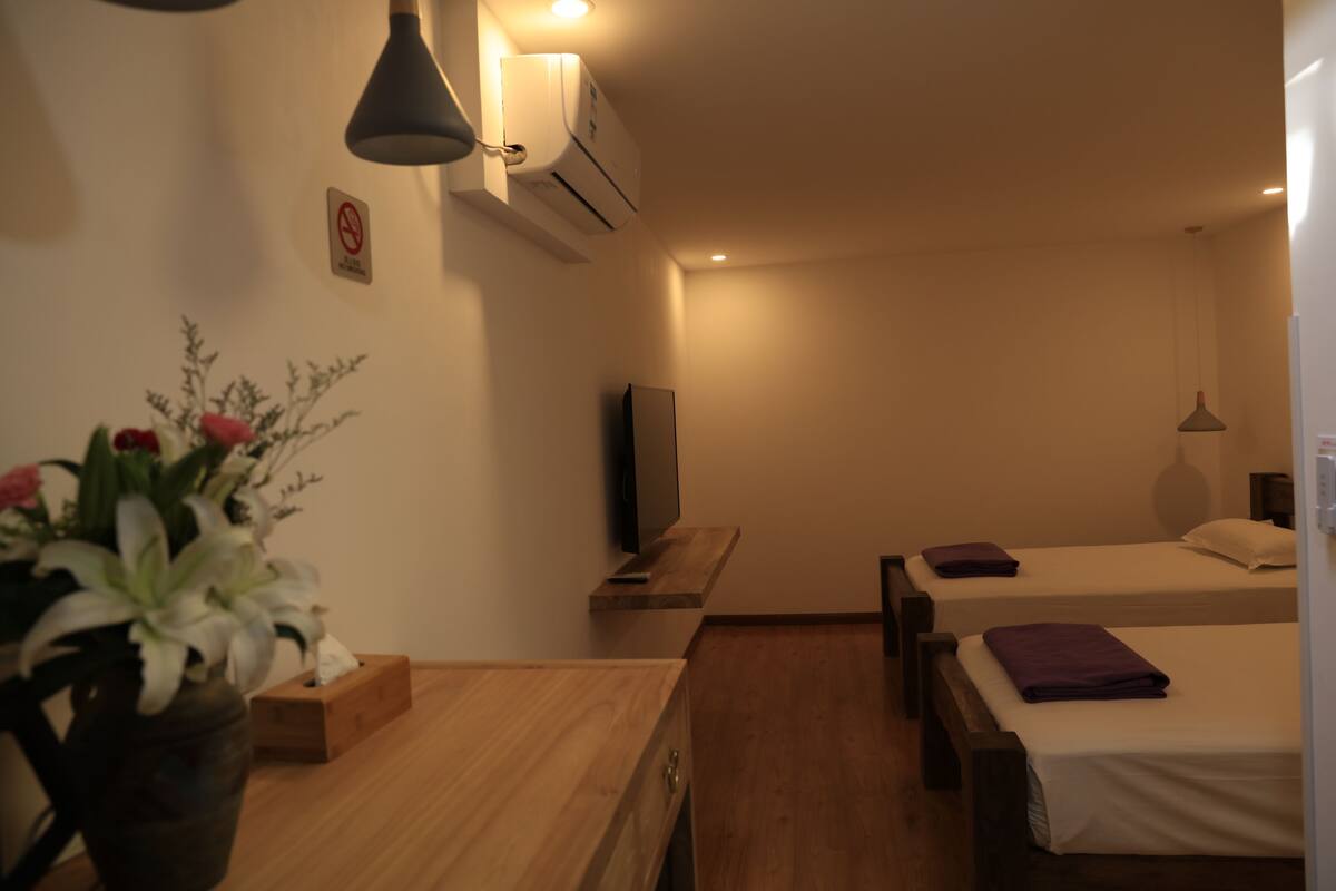 A hotel room with two beds, a wooden desk, a TV, and a wall-mounted air conditioner.