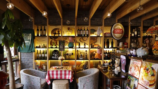 A cozy bar area with wooden beams, shelves of bottles, and a variety of bar tools. There are two upholstered chairs and a table with a checkered tablecloth. - Beijing Downtown Travelotel (Beijing)
