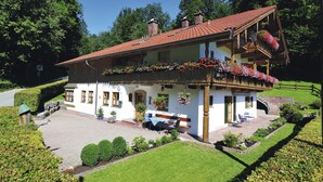 A two-story chalet with a red-tiled roof, a balcony with hanging flower baskets, and a paved courtyard with seating and a small table. - Gaestehaus Vogl (Berchtesgaden)