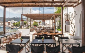 An outdoor dining area with wooden tables and chairs, potted plants, and hanging lamps. - CHRIS HOTEL (Patmos)