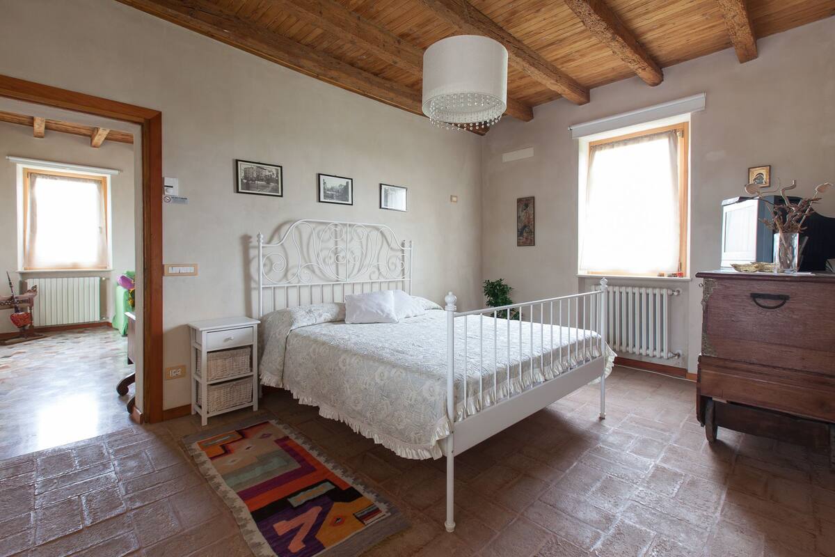 A bedroom with a white bed, a wooden ceiling, a white radiator, and a window with a curtain.