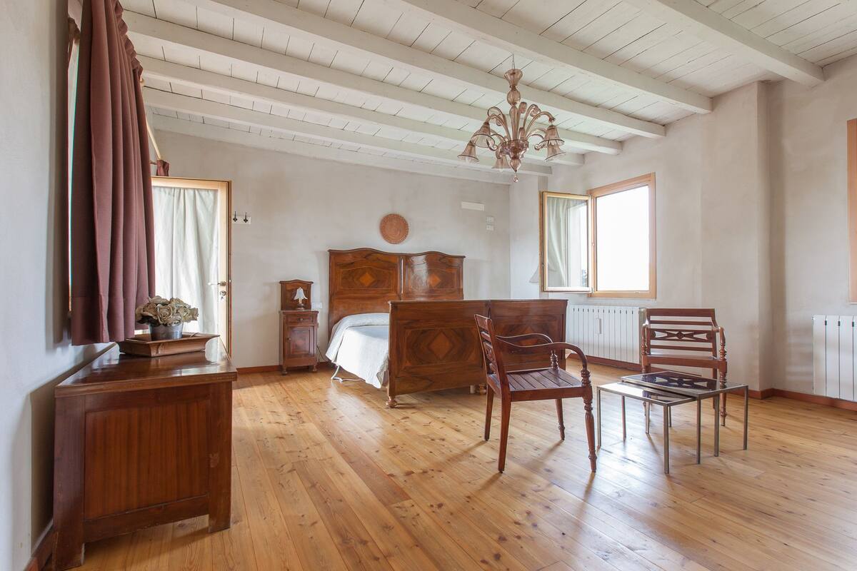 A bedroom with a wooden bed, a wooden chair, a small table, a nightstand, a chandelier, and a window.