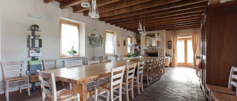 Un comedor amplio con una larga mesa de madera y sillas blancas, que cuenta con un techo rústico y suelo de piedra.