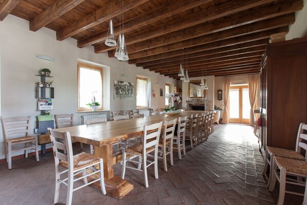 A spacious dining area with a long wooden table and white chairs, featuring a rustic ceiling and stone flooring.