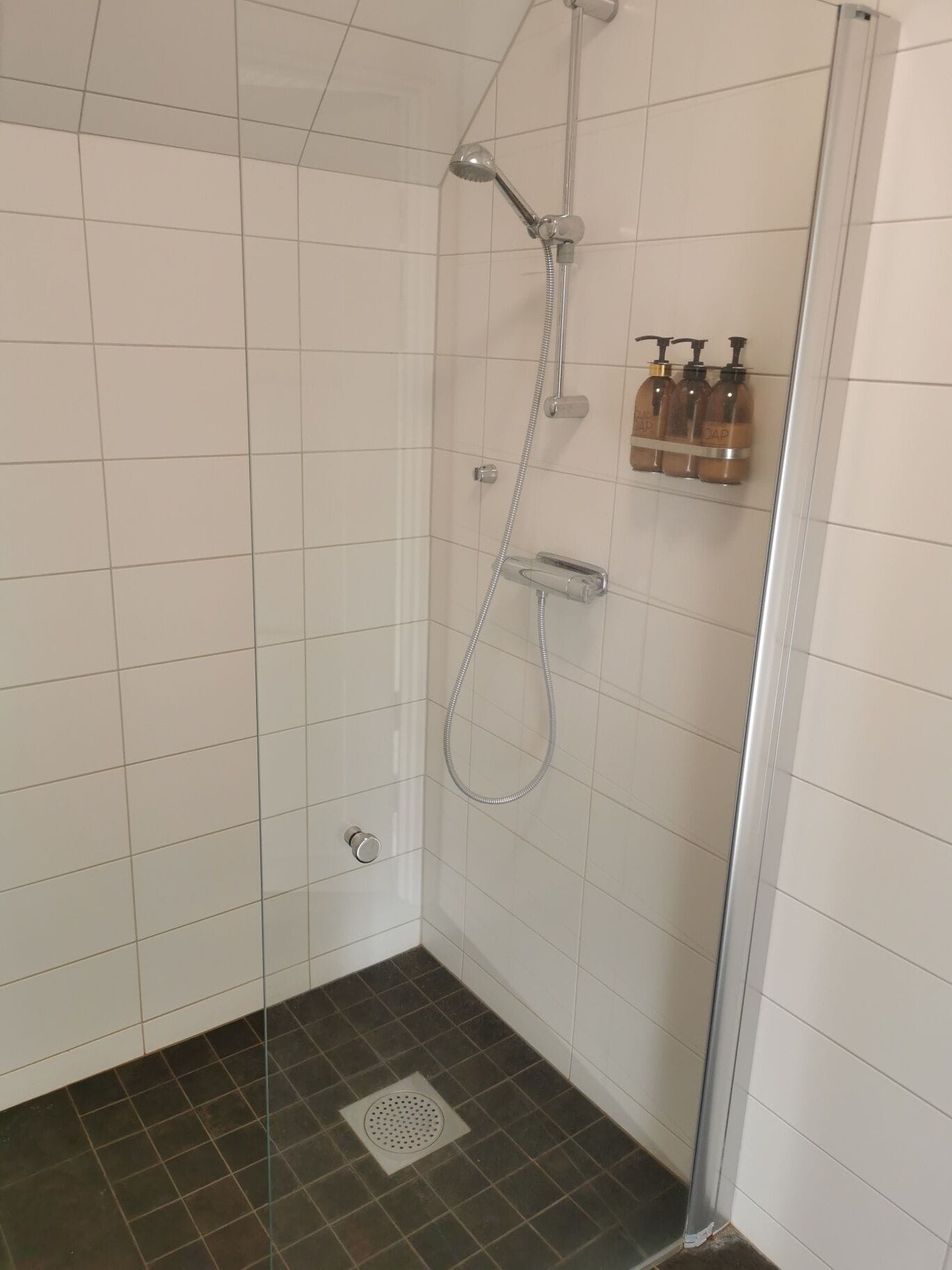 A shower with a handheld showerhead and a wall-mounted faucet. There are three bottles of soap on the wall.