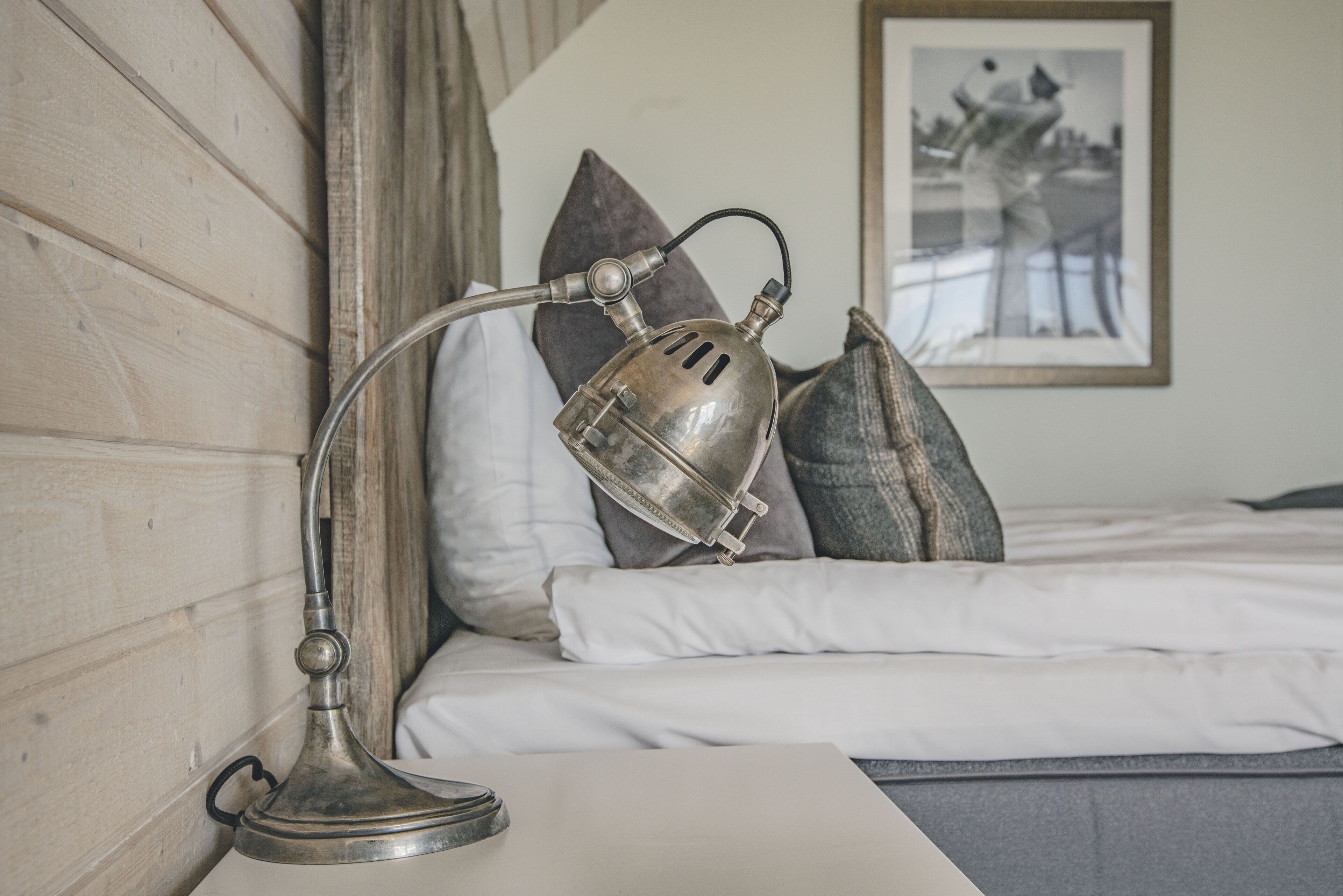 A bed with white linens, a metal lamp on a bedside table, and a framed photograph on the wall.