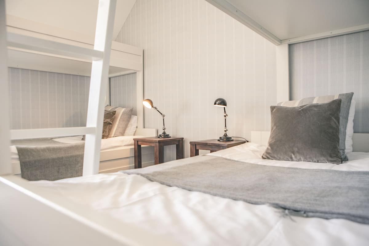 Ett modernt hotellrum med en våningssäng, ett sängbord med en lampa, ett litet sidobord och en stor säng med ett grått överkast.