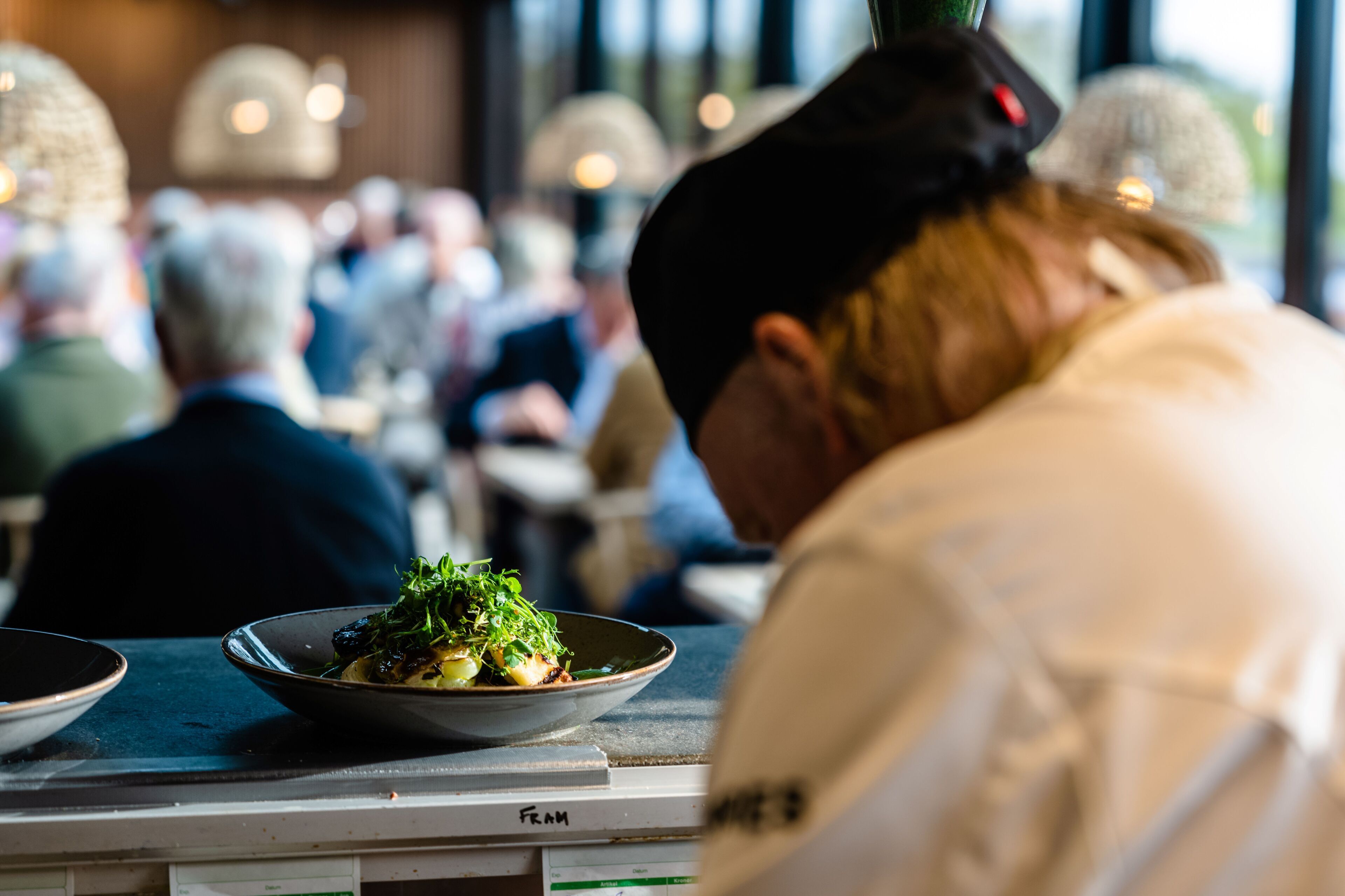 A restaurant interior with diners seated at tables, a chef in the foreground, and a dish on a table.