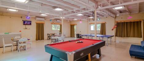 A pool table, a ping pong table, and a billiards table in a well-lit indoor space with ceiling lights and windows.
