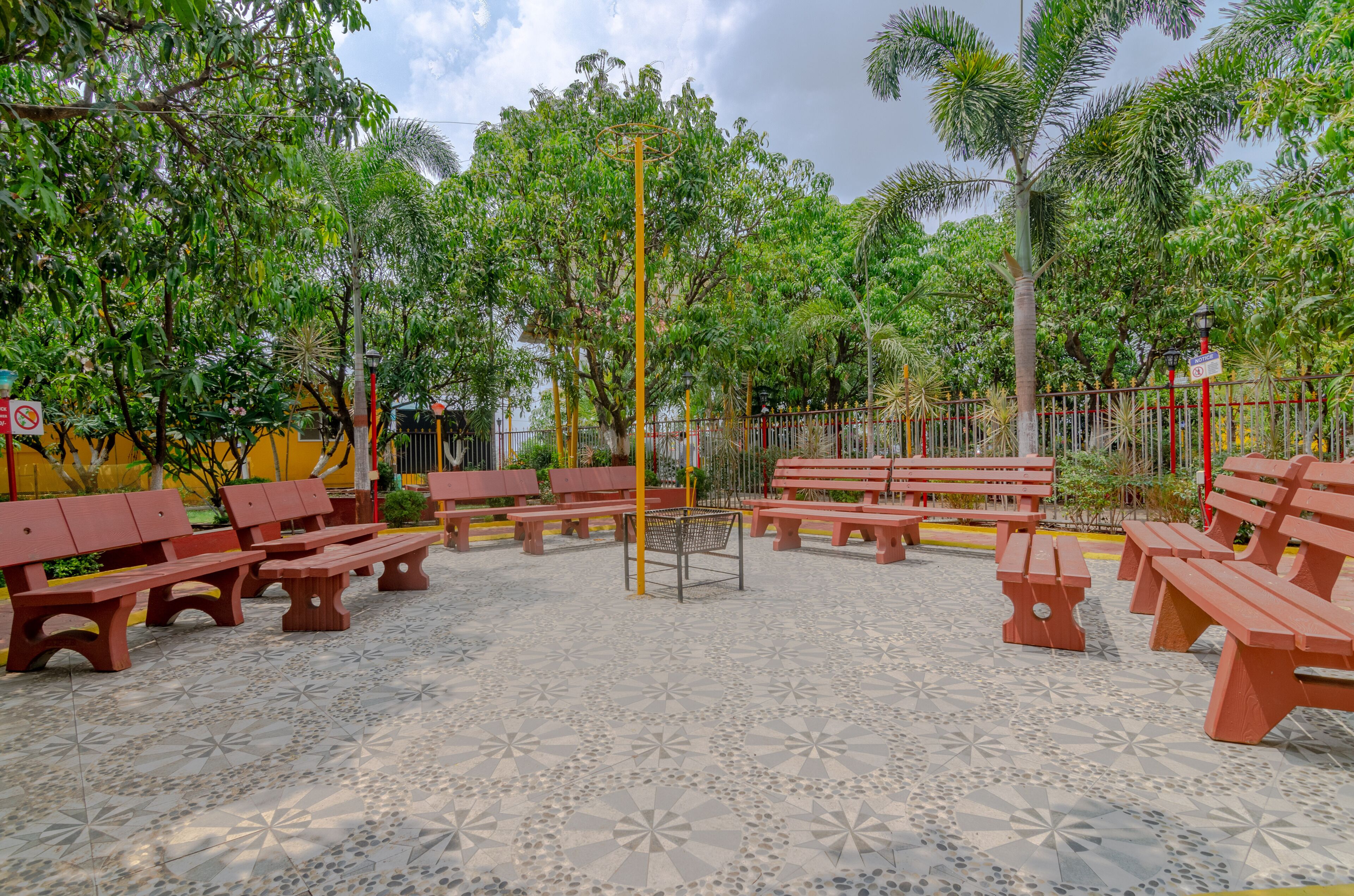 Un patio con bancos rojos dispuestos en semicírculo, rodeado de árboles y un suelo de baldosas.