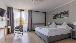 A modern hotel room with a large bed, a desk with a chair, a TV, and a window with curtains. - Bildungs- und Tagungshaus Haus Neuland (Bielefeld)