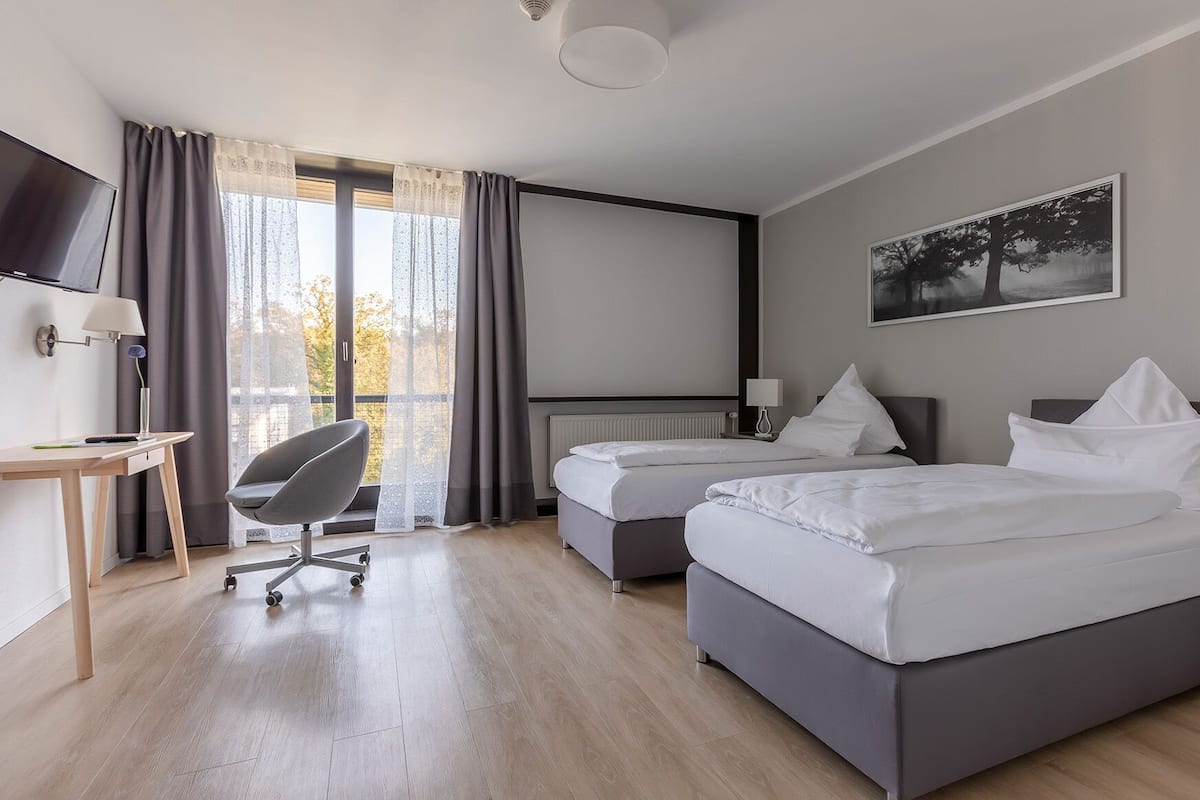 A hotel room with two beds, a desk, a chair, a TV, and a window with curtains.