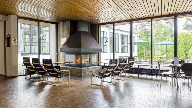 A spacious indoor area with a large fireplace, wooden ceiling, and large windows offering a view of the outdoors. - Bildungs- und Tagungshaus Haus Neuland (Bielefeld)