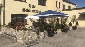 Um hotel chamado "Hotel Schützenhaus" com área externa para sentar, vasos de pedra com flores e um céu azul claro.