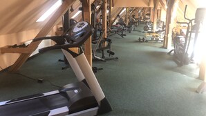 Academia com diversos equipamentos de exercício, incluindo esteiras e máquinas de musculação, num espaço com vigas de madeira e teto inclinado.