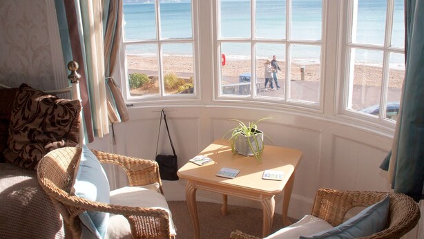 A room with a large bay window overlooking a beach, wicker furniture, and a small table with a plant. - Lyndale Guest House (Weymouth)