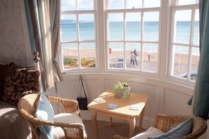 A room with a large bay window overlooking a beach, wicker furniture, and a small table with a plant. - Lyndale Guest House (Weymouth)