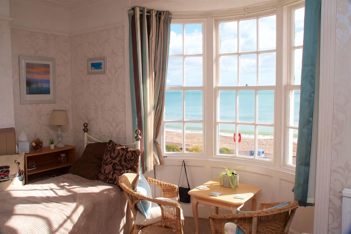 A bedroom with a bed, a chair, a table, and a view of the beach through large windows.