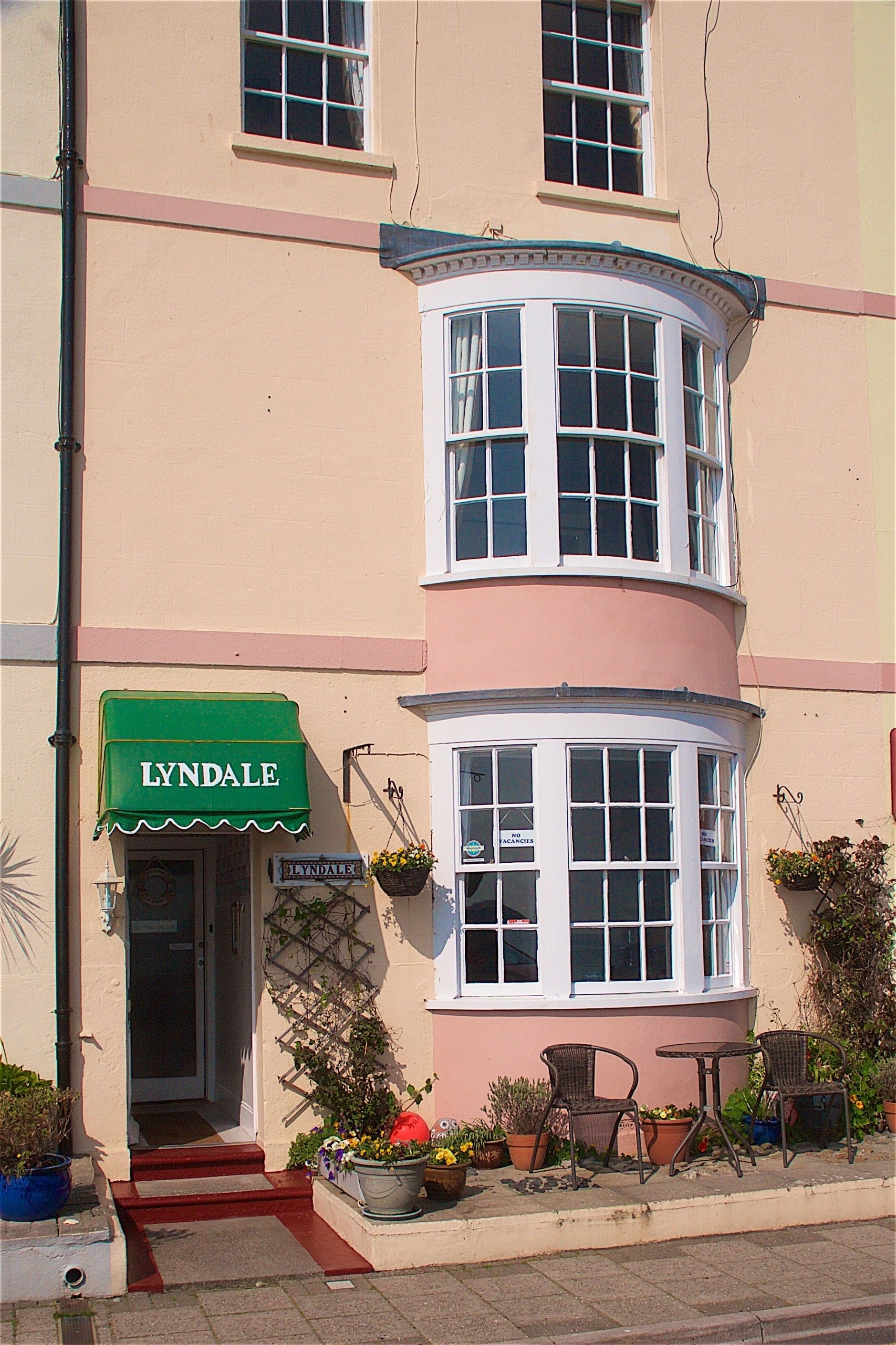 Un edificio en esquina con ventanal saliente, toldo verde con la palabra 'LYNDALE' escrita y mesas y sillas para terraza.