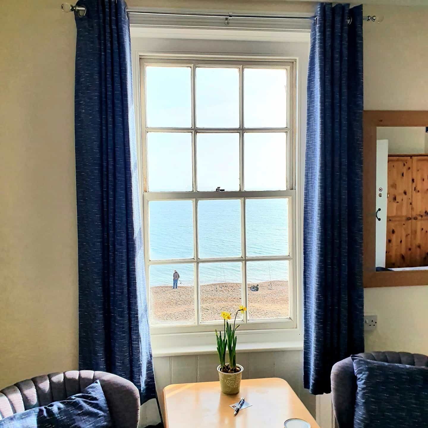 A room with a large window overlooking a beach, a small table with a plant, and two chairs.