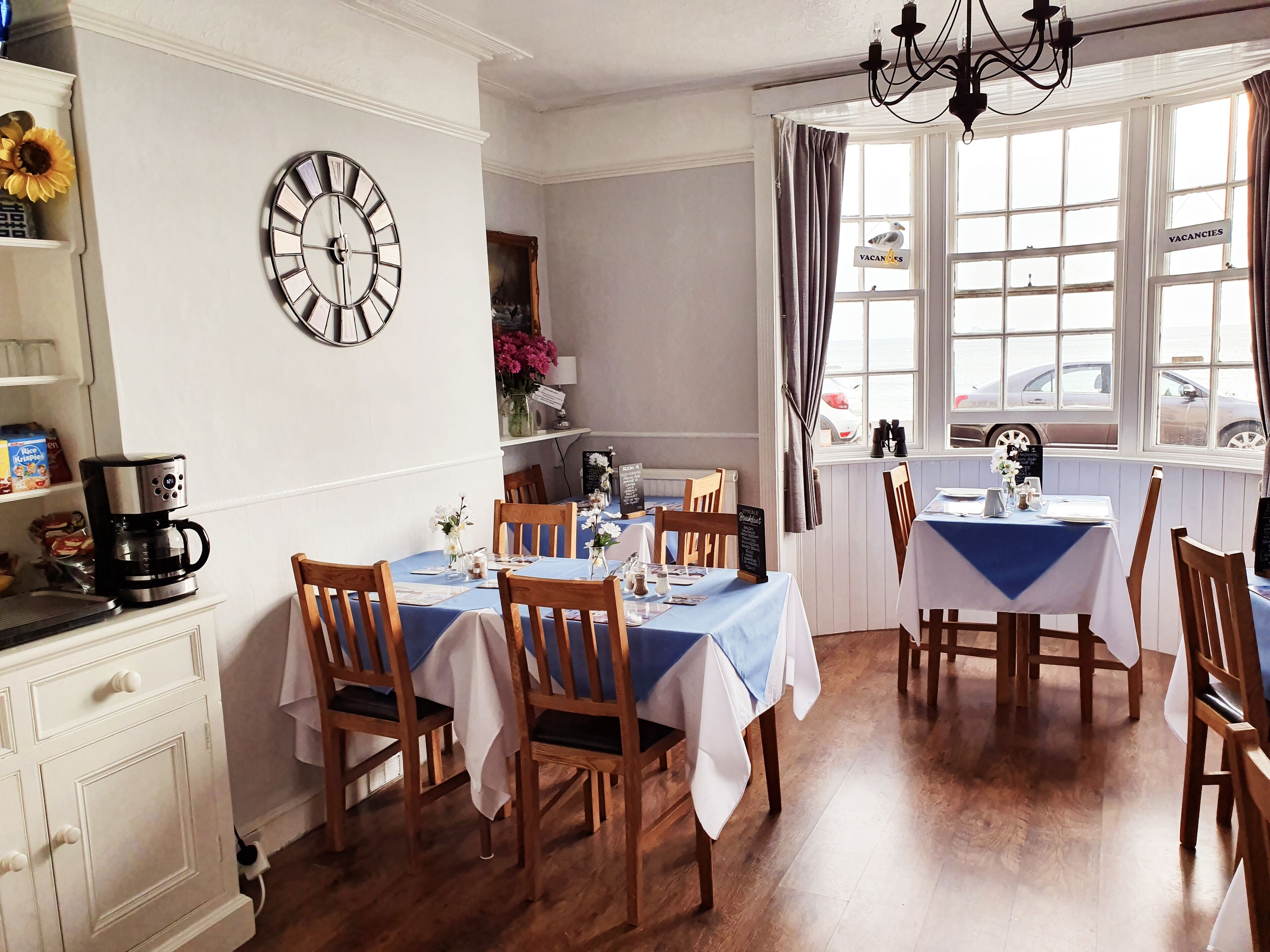 A dining area with wooden tables and chairs, a clock on the wall, and a view of the outside through large windows.