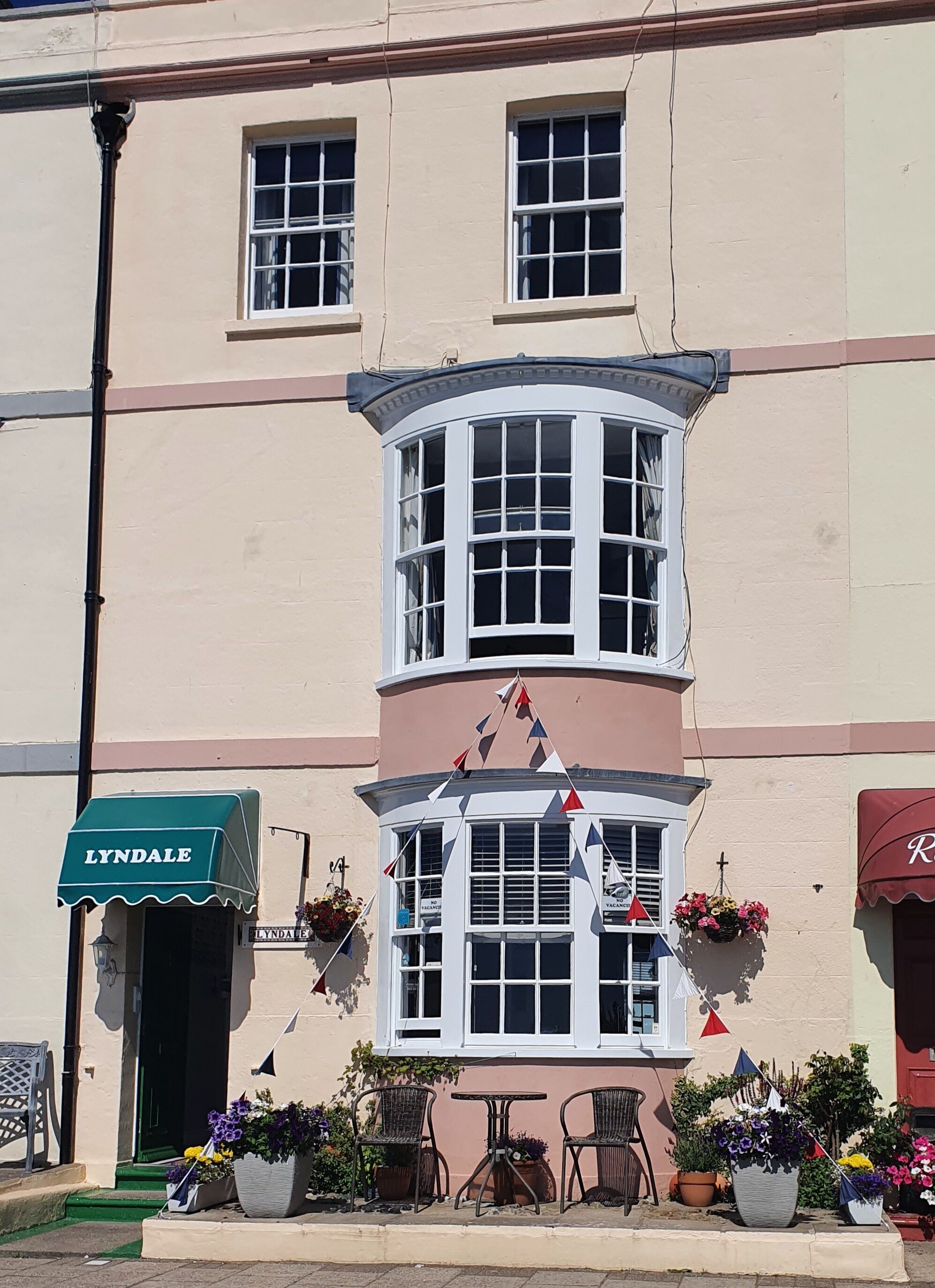 A corner building with a bay window, a green awning labeled 'LYNDAL', outdoor seating, and flower pots.