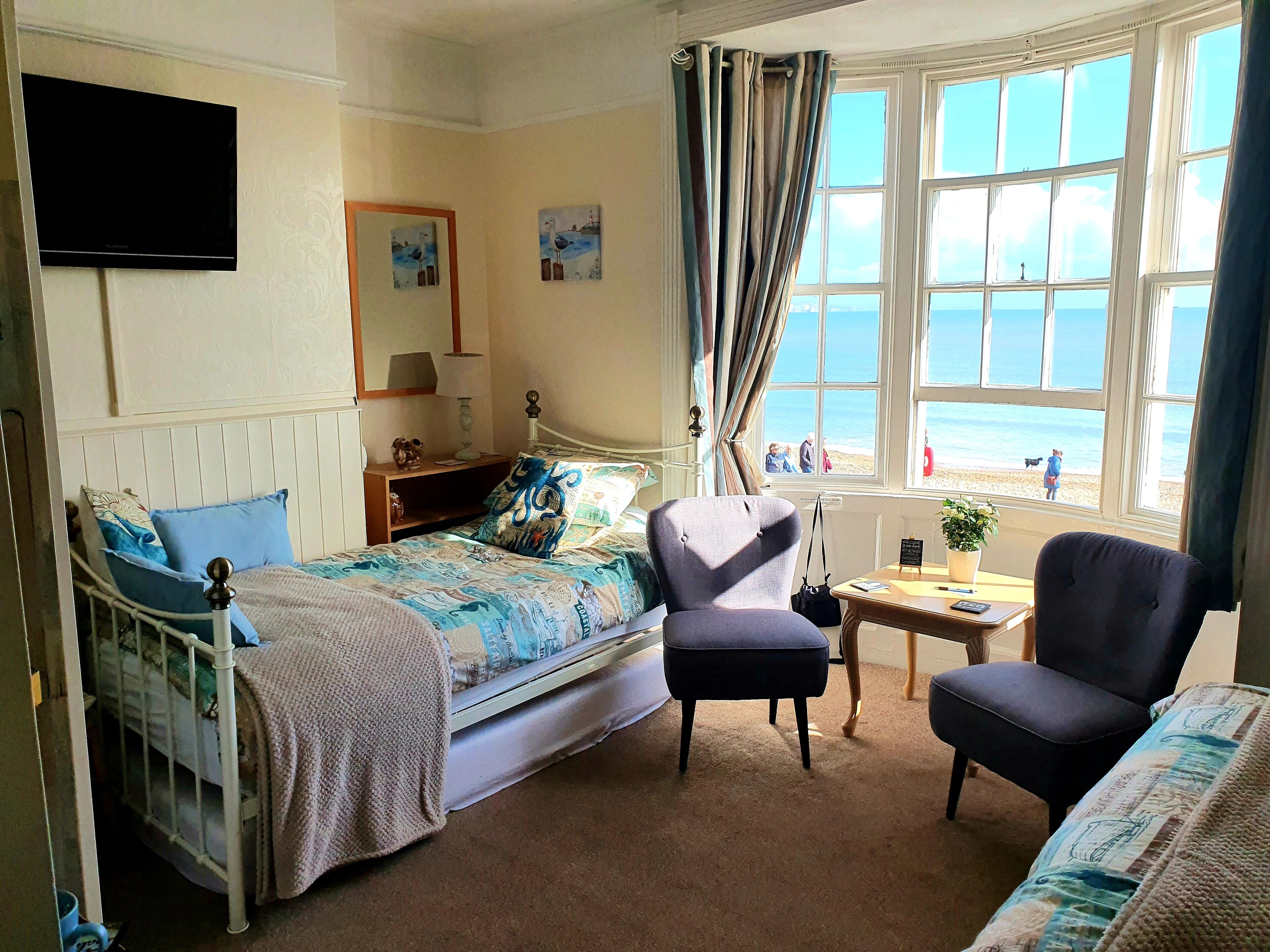 A bedroom with a bed, two chairs, a small table, a TV, and a window with a view of the beach.