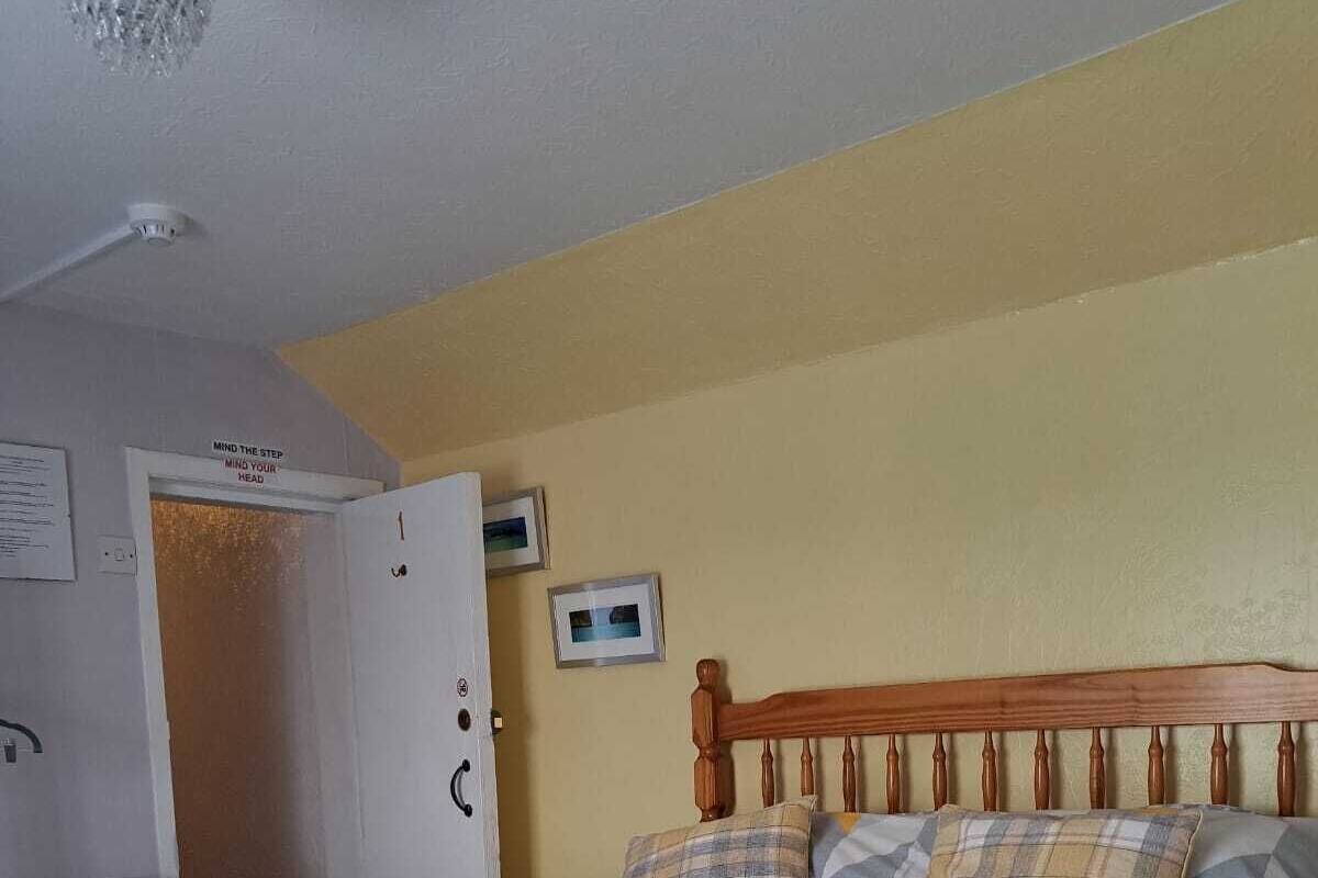 A bedroom with a bed, a chair, a door, and a wall-mounted thermostat.