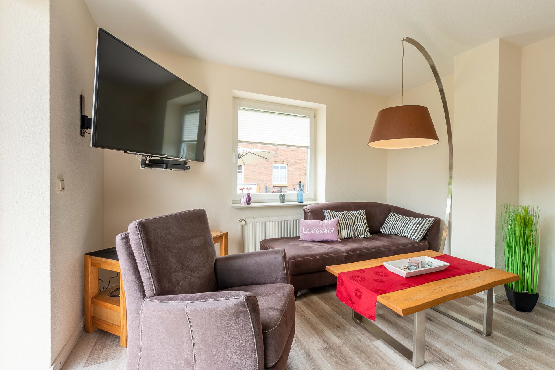 A modern living room with a sofa, a chair, a TV mounted on the wall, a table with a red tablecloth, and a potted plant.