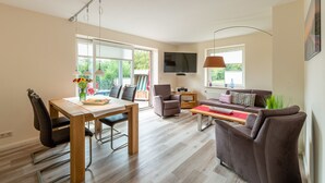 A modern living room with a dining area, a flat-screen TV mounted on the wall, and large windows providing natural light. - Haus Bendix (Dagebuell)