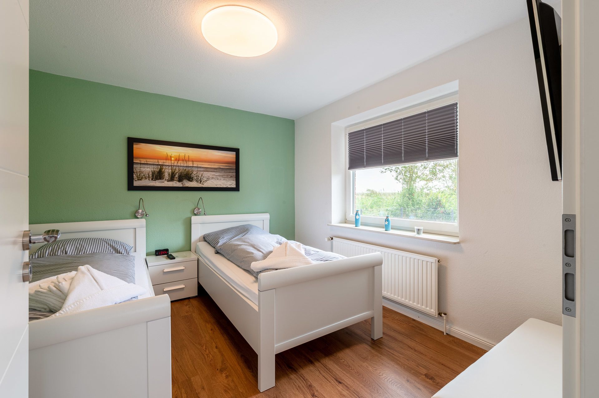 Une chambre à coucher comprenant un lit, des tables de chevet, une commode, une fenêtre avec des stores, un radiateur et un tableau accroché au mur.