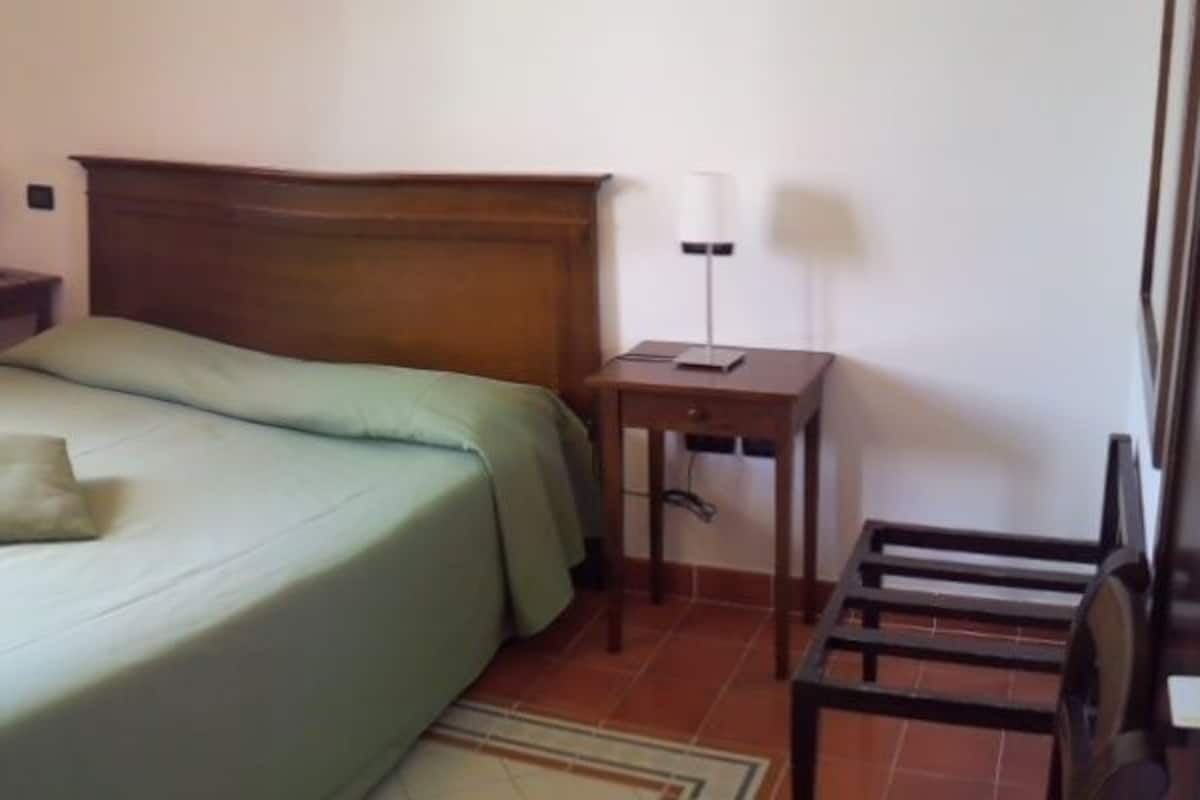 Um quarto com cama, mesa de cabeceira, cadeira e janela.